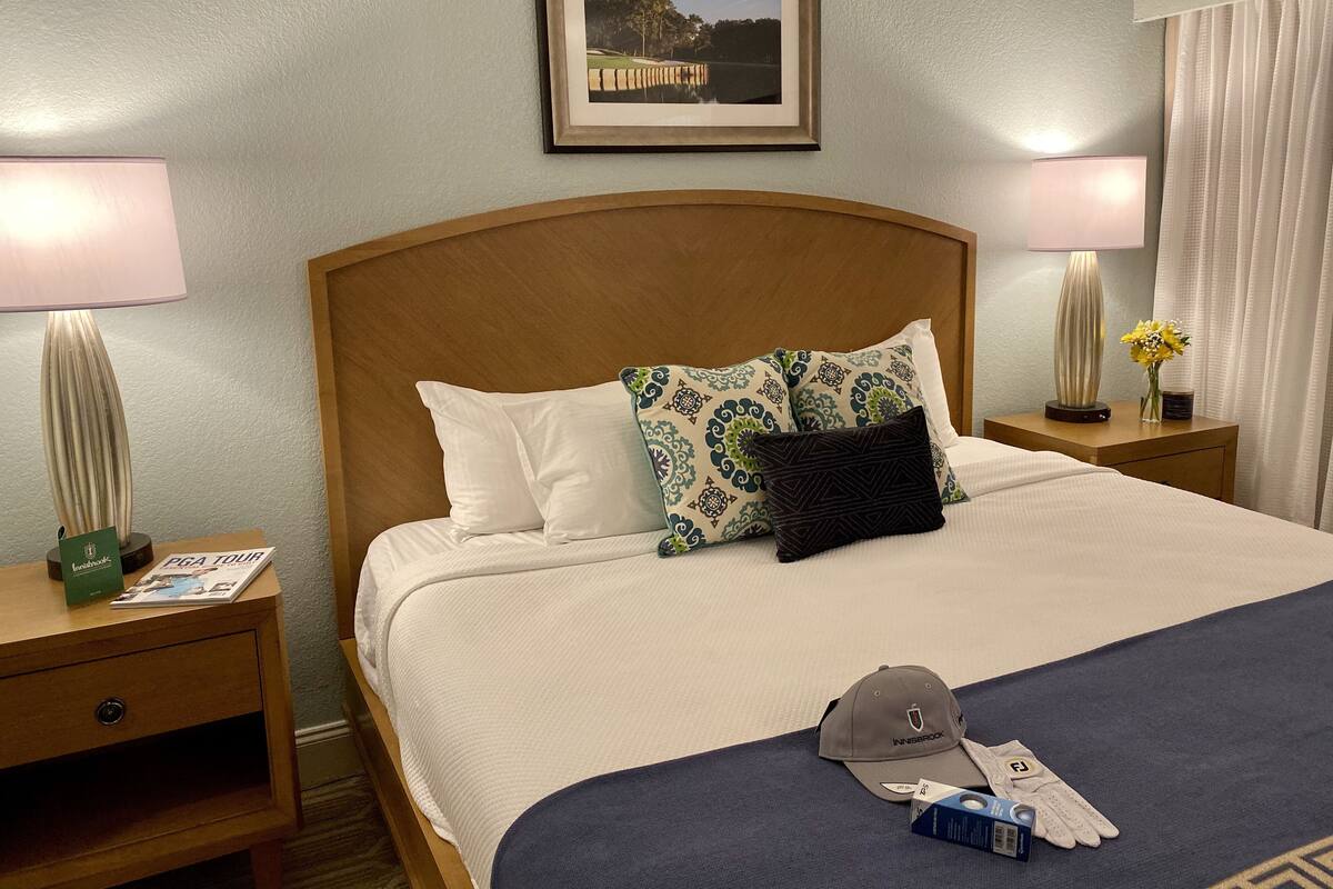A neatly made bed with a headboard, two bedside tables with lamps, a framed picture on the wall, and a small table with a vase of flowers.
