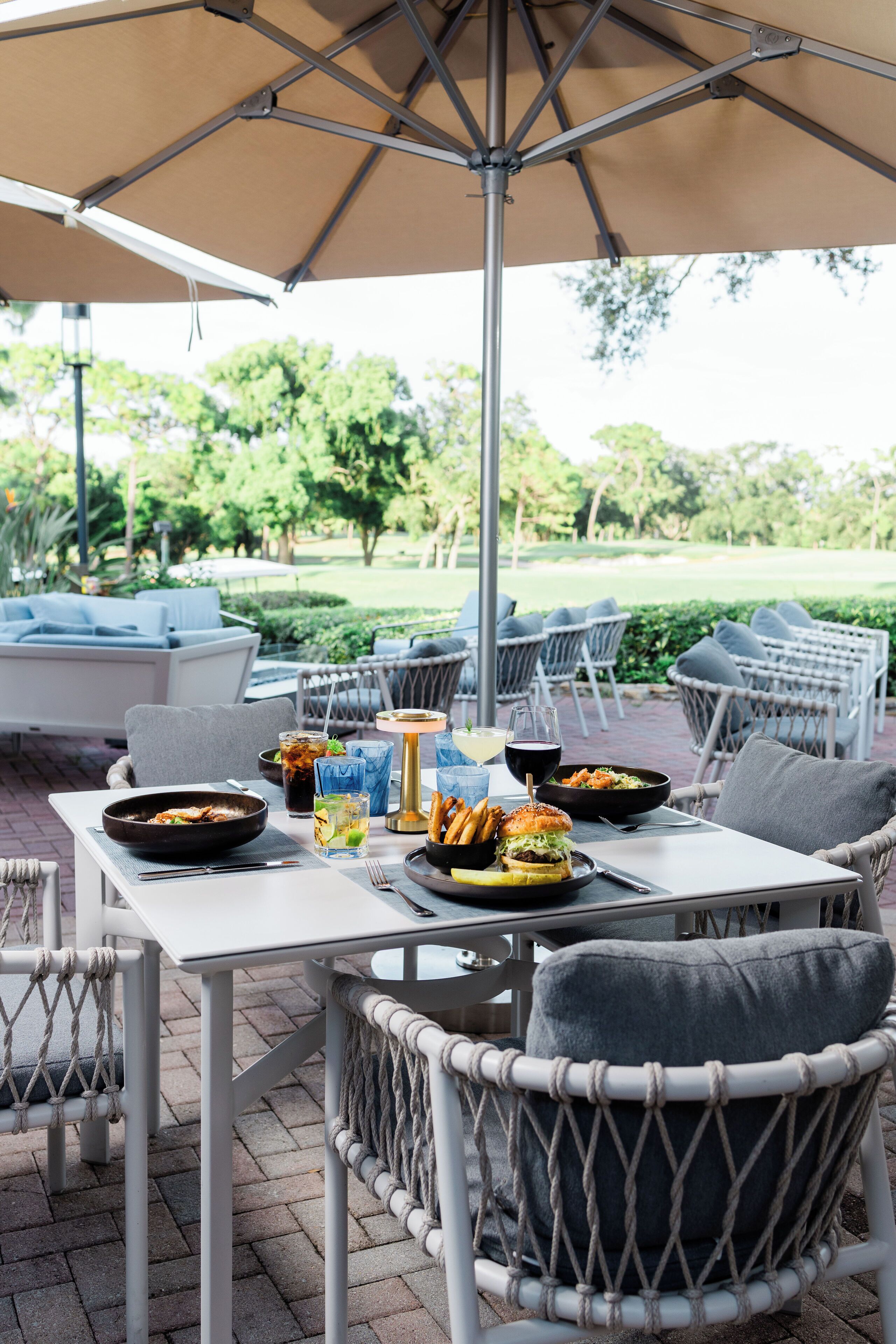 An outdoor dining area with wicker chairs, a table with food and drinks, and a large umbrella providing shade.
