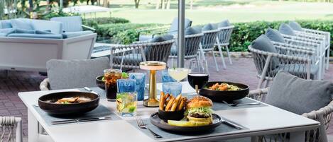 An outdoor dining area with wicker chairs, a table with food and drinks, and a large umbrella providing shade.