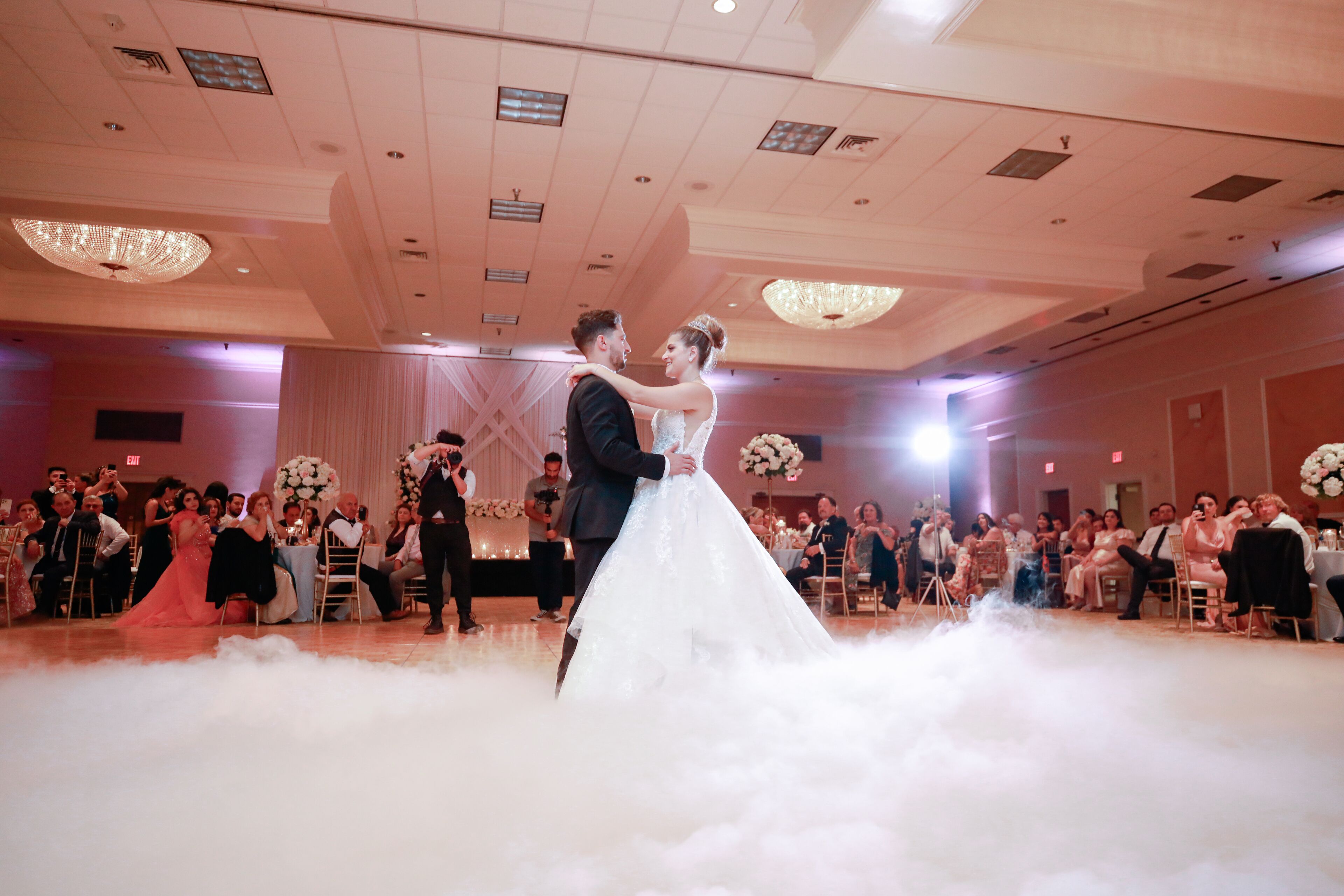 A wedding reception with a couple dancing on a cloud-like floor effect, surrounded by guests seated at round tables with floral centerpieces.