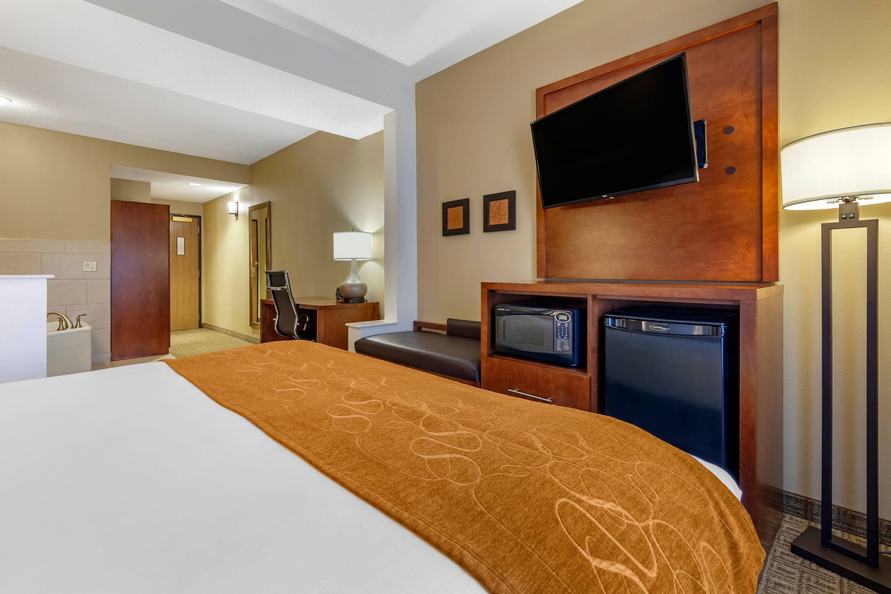 A hotel room with a large bed, a flat-screen TV mounted on the wall, a microwave, a mini fridge, a desk with a lamp, and a bathroom visible in the background.
