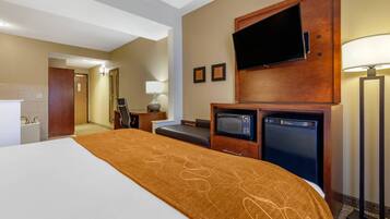 A hotel room with a large bed, a flat-screen TV mounted on the wall, a microwave, a mini fridge, a desk with a lamp, and a bathroom visible in the background.
