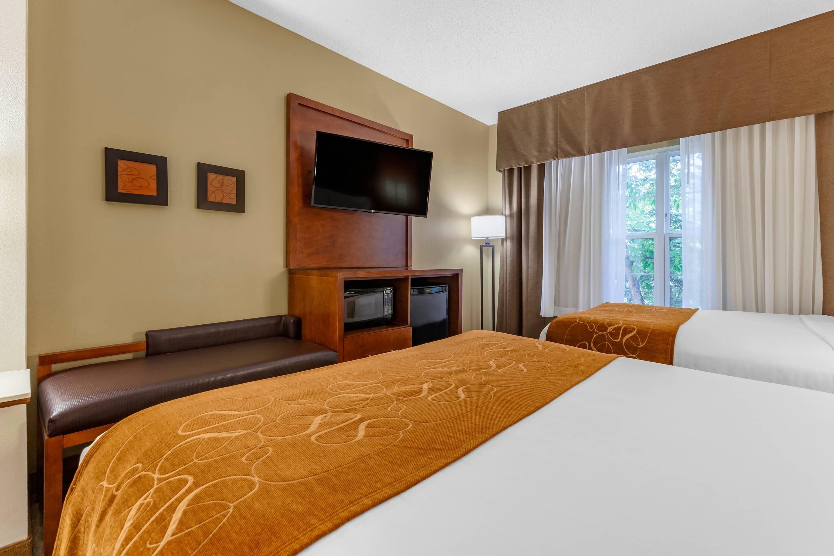 A hotel room with a large bed, a TV mounted on a wooden panel, a small seating area, and a window with curtains.