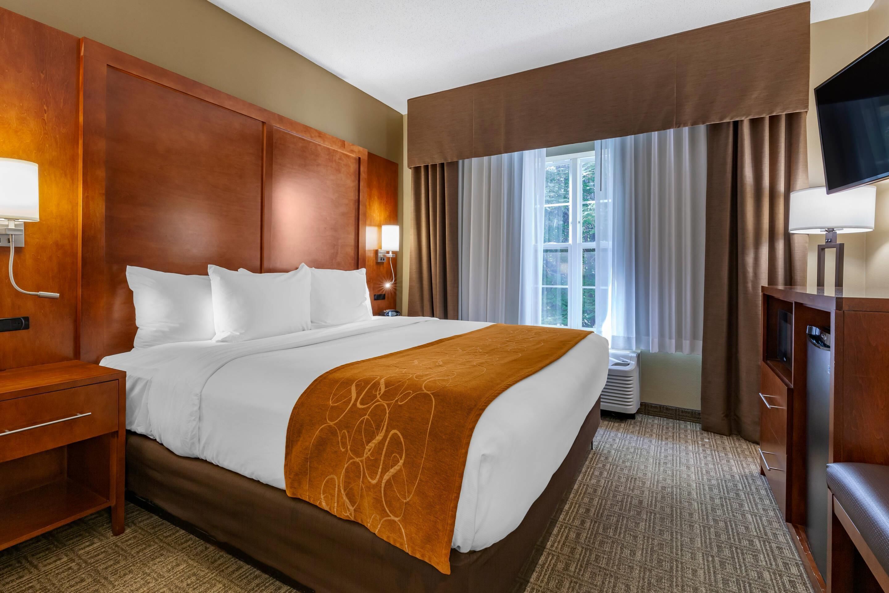Une chambre d’hôtel avec un grand lit, une tête de lit en bois, un téléviseur à écran plat, un bureau et une fenêtre avec des rideaux.