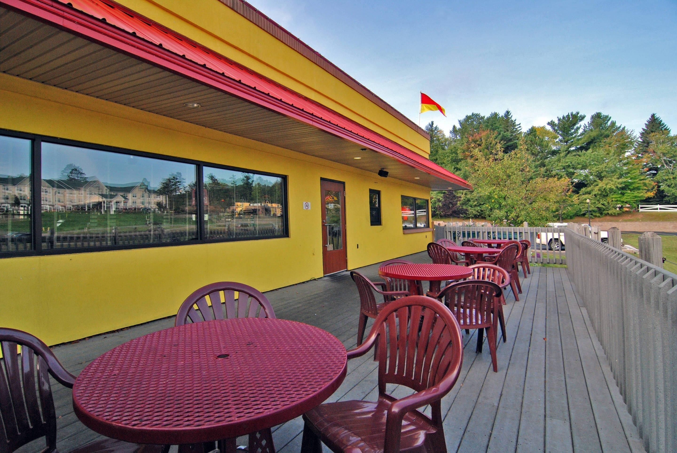 Un bâtiment jaune avec un auvent rouge et un drapeau, des tables et des chaises à l’extérieur, et une vue sur un terrain de sport.