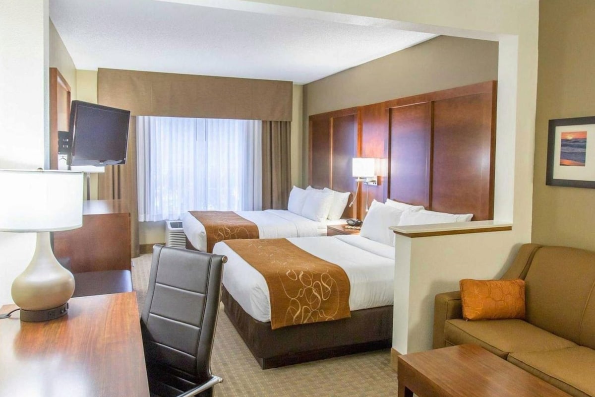 A hotel room with two beds, a desk, a chair, a sofa, a TV, and a window with curtains.