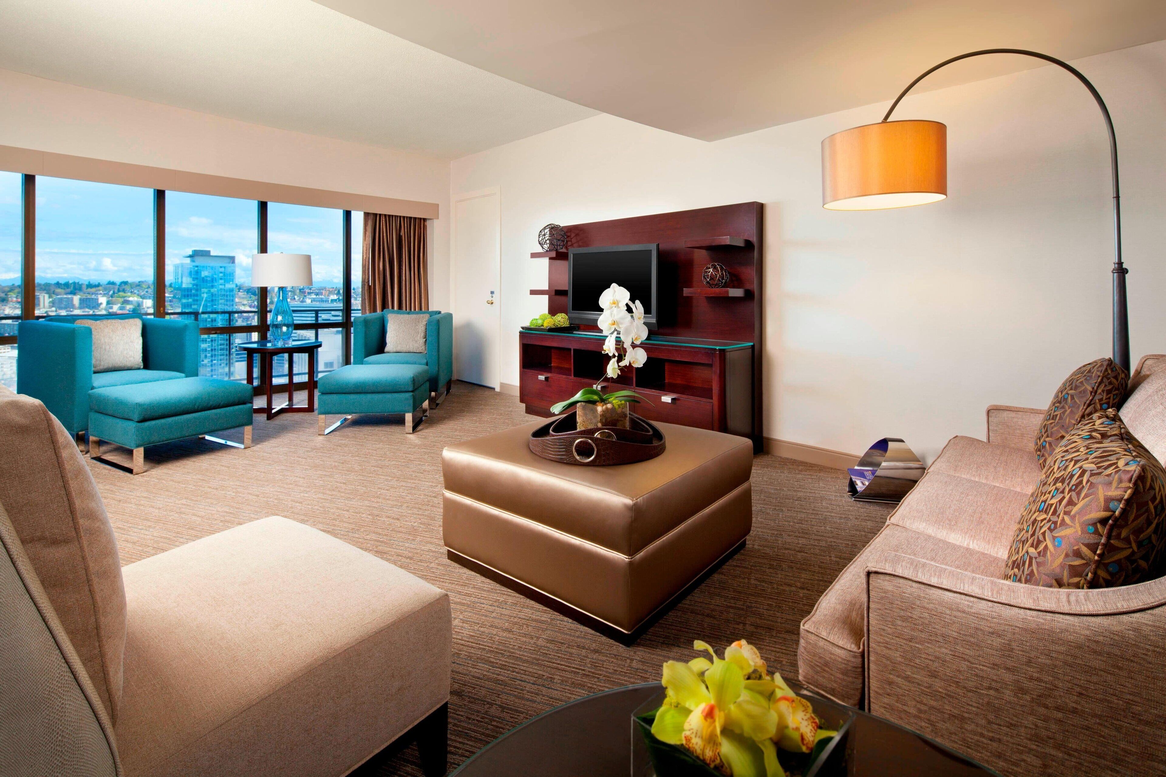 A modern hotel room with a large window offering a city view, a brown leather sofa, a teal armchair, a small coffee table, and a TV on a wooden stand.