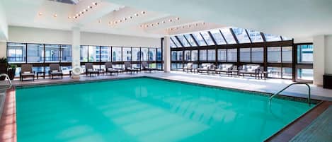 Une piscine intérieure à l’eau d’un bleu cristallin, entourée de chaises longues et de tables, de grandes fenêtres offrant une vue sur la ville et un design de plafond moderne.