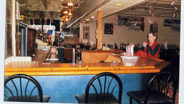A bar with a counter, chairs, and a person seated at a table. - Will Rogers Inn (Claremore)
