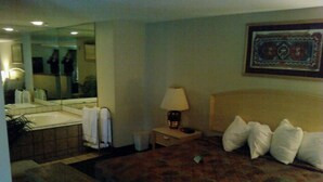 A hotel room with a bed, a nightstand with a lamp, a mirror, a bathroom with a sink and a bathtub. - Will Rogers Inn (Claremore)