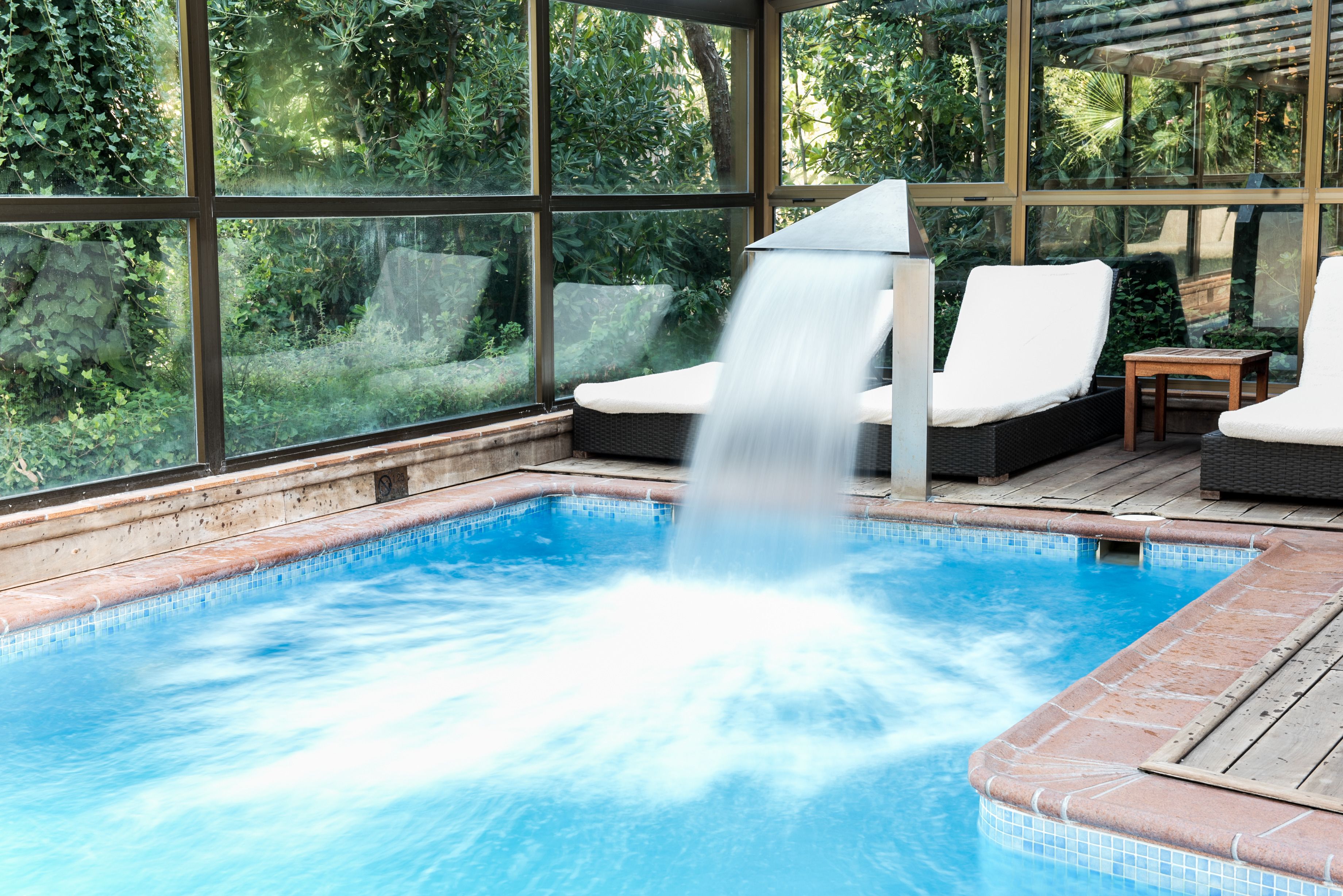 A hot tub with a waterfall feature, surrounded by large windows and outdoor furniture.