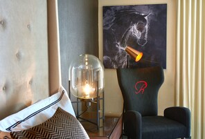A modern hotel room with a tufted headboard, a clear glass lamp, a black chair with a red logo, and a large abstract painting on the wall.