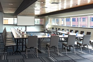 A conference room with a long table, chairs, a projector screen, and windows with a view of buildings.