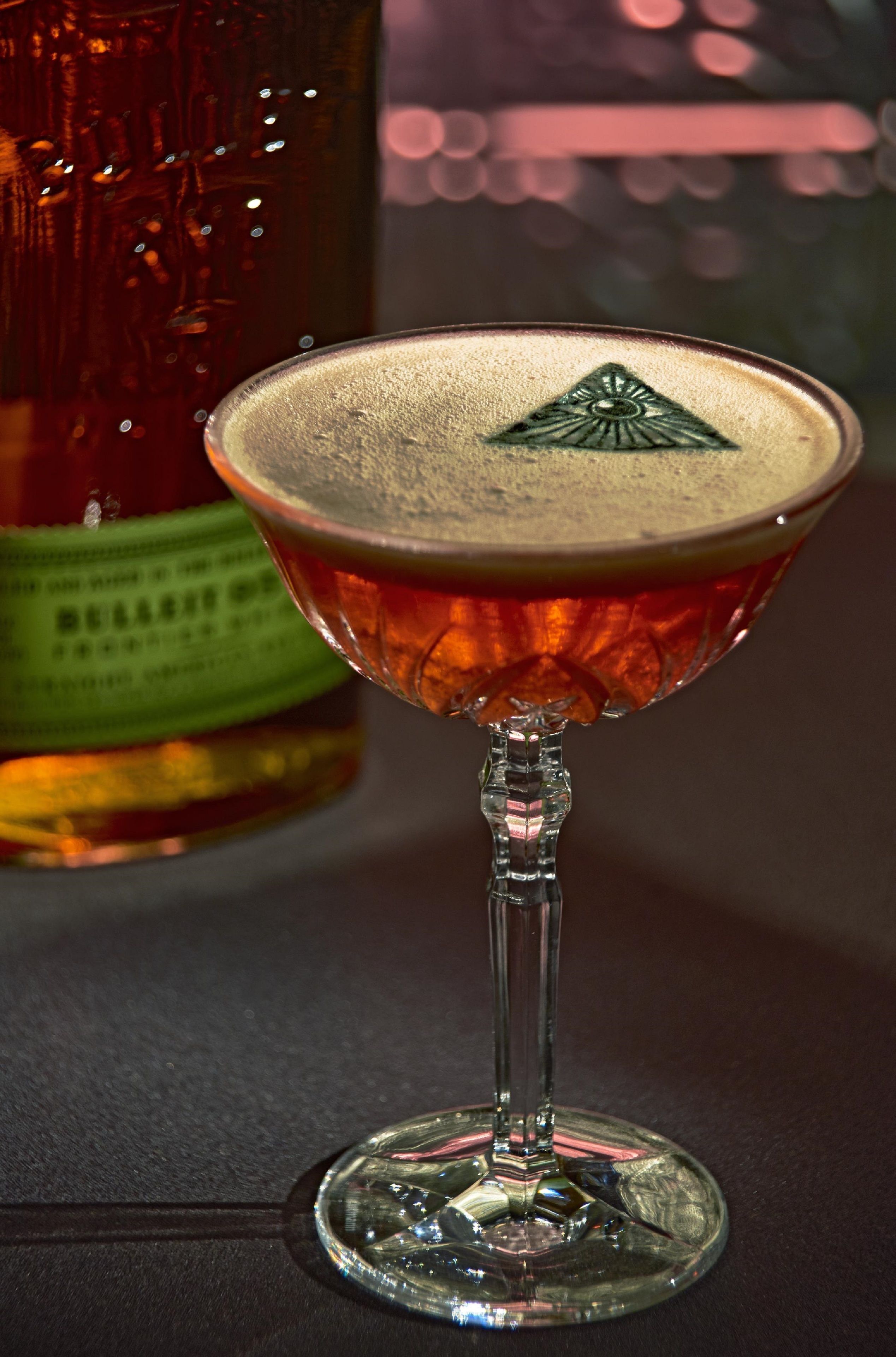 A cocktail in a martini glass with a decorative emblem on the rim, placed on a table with a bottle in the background.