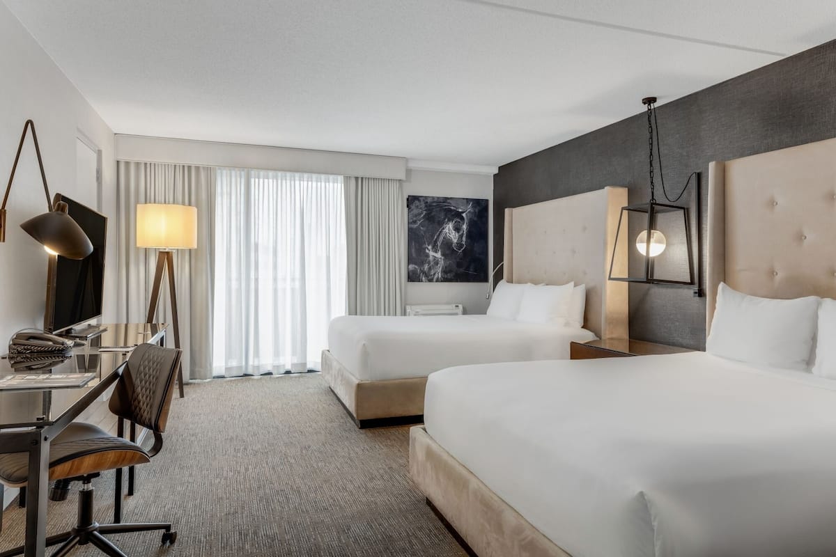 Ein Hotelzimmer mit zwei Betten, einem Schreibtisch mit Computer, einem Stuhl, einer Lampe und einem großen Fenster mit Vorhängen.