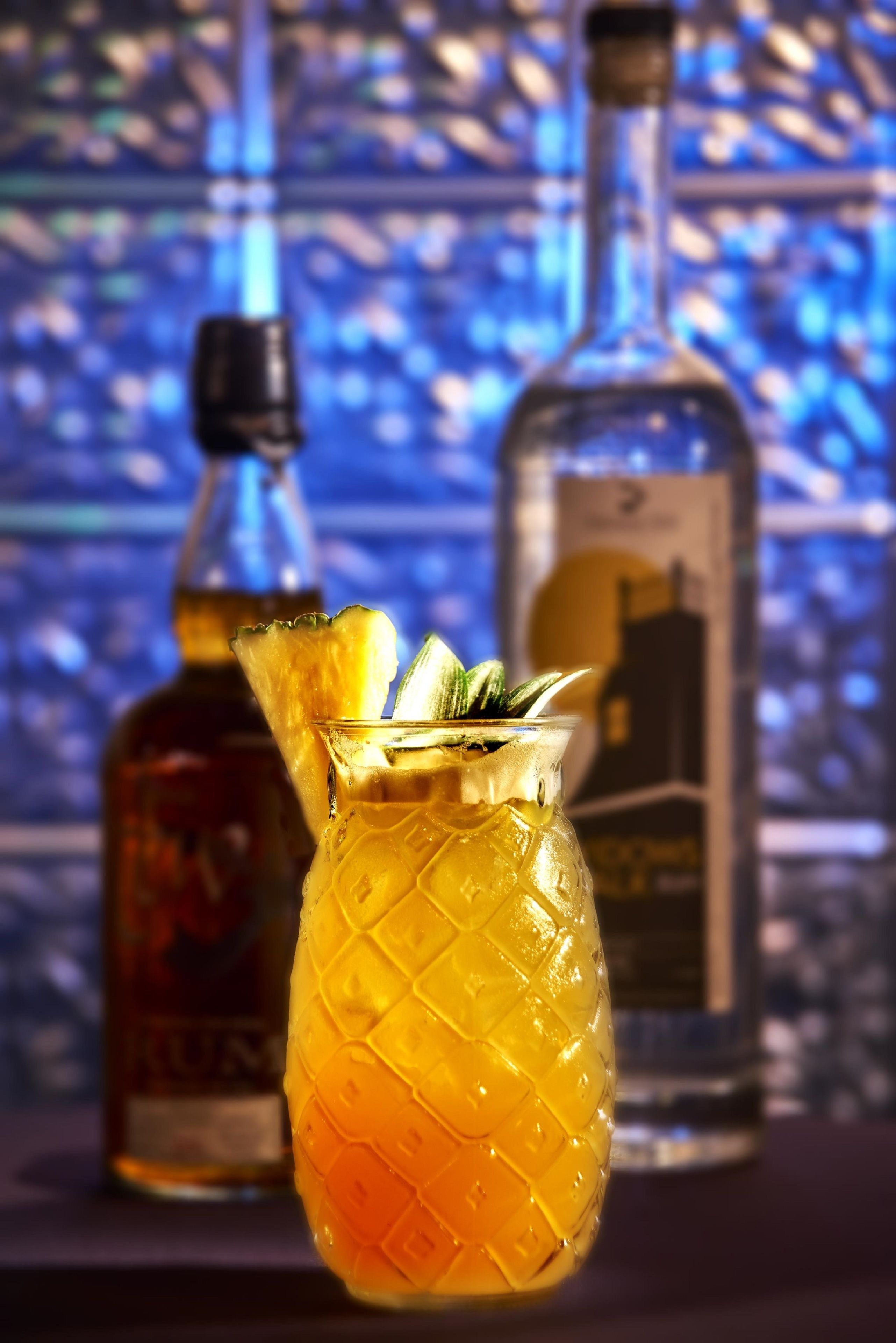En cocktail i ett glas formad som en ananas, med en flaska sprit i bakgrunden.