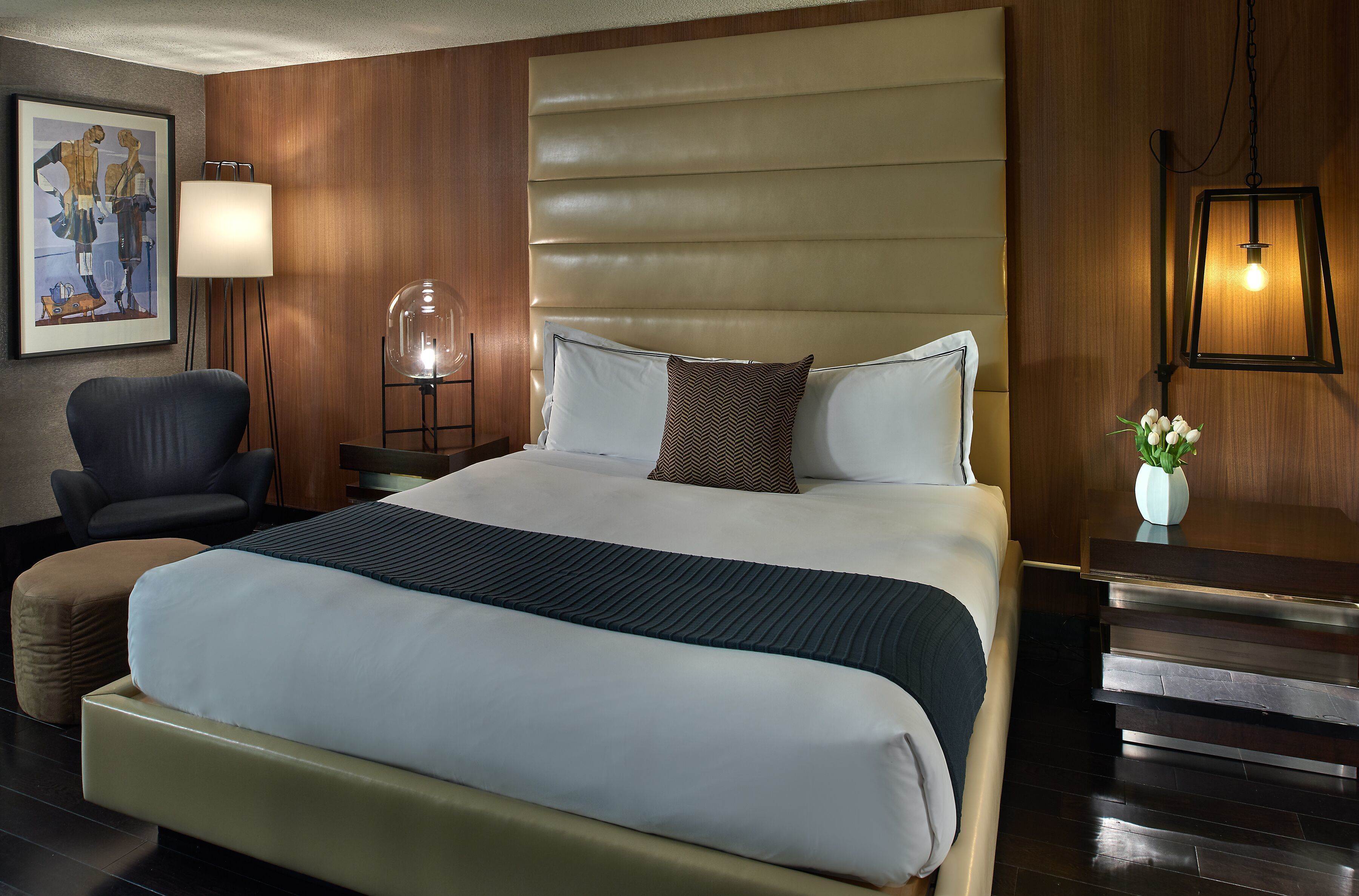 Quarto de hotel com uma cama grande, uma cadeira, uma mesinha de cabeceira, uma luminária e um quadro na parede.