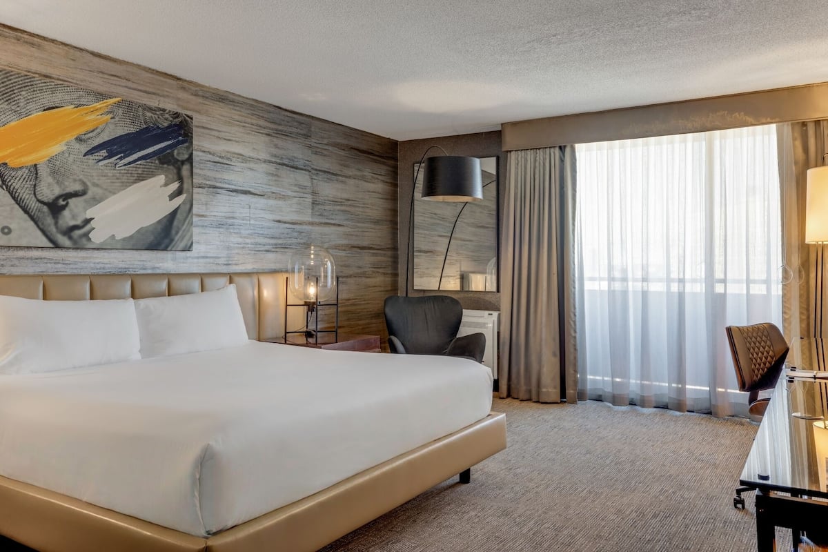 Ein Hotelzimmer mit einem großen Bett, einem Schreibtisch, einem Stuhl, einer Lampe und einem fenster mit Vorhängen.