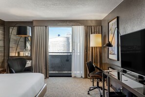 A hotel room with a bed, desk, chair, TV, and a view of a cityscape through the window.