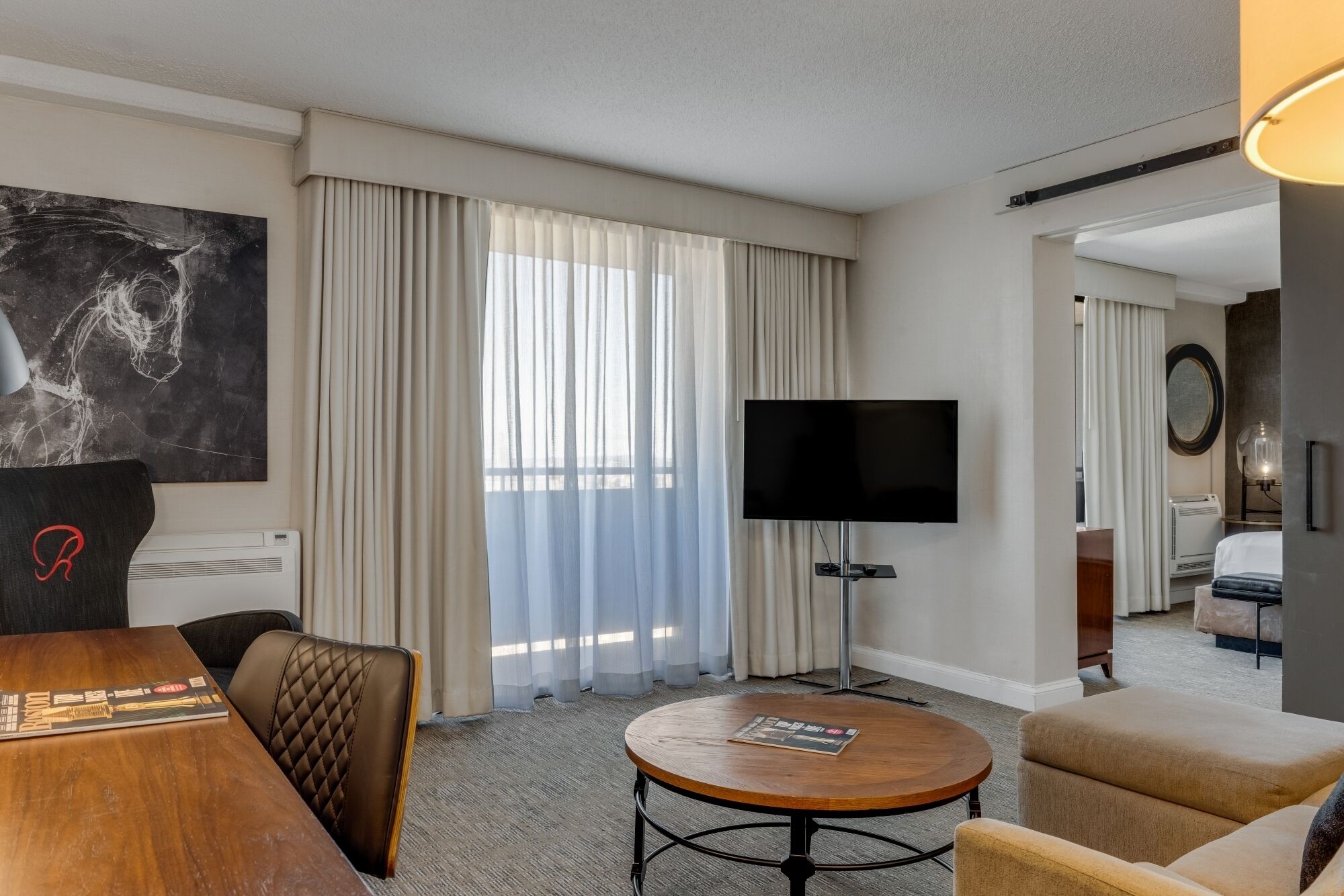 A hotel room with a flat-screen TV, a round wooden table, a sofa, a bed, and a large window with curtains.
