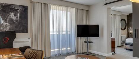 A hotel room with a flat-screen TV, a round wooden table, a sofa, a bed, and a large window with curtains.