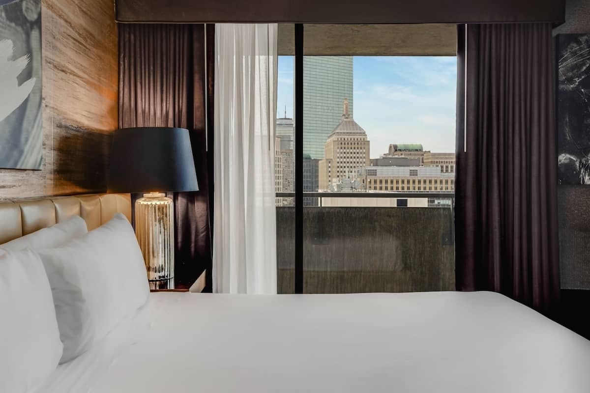 Ein Hotelzimmer mit Bett, Lampe und Blick auf Stadtgebäude durch große Fenster, sowie einem Vorhang.