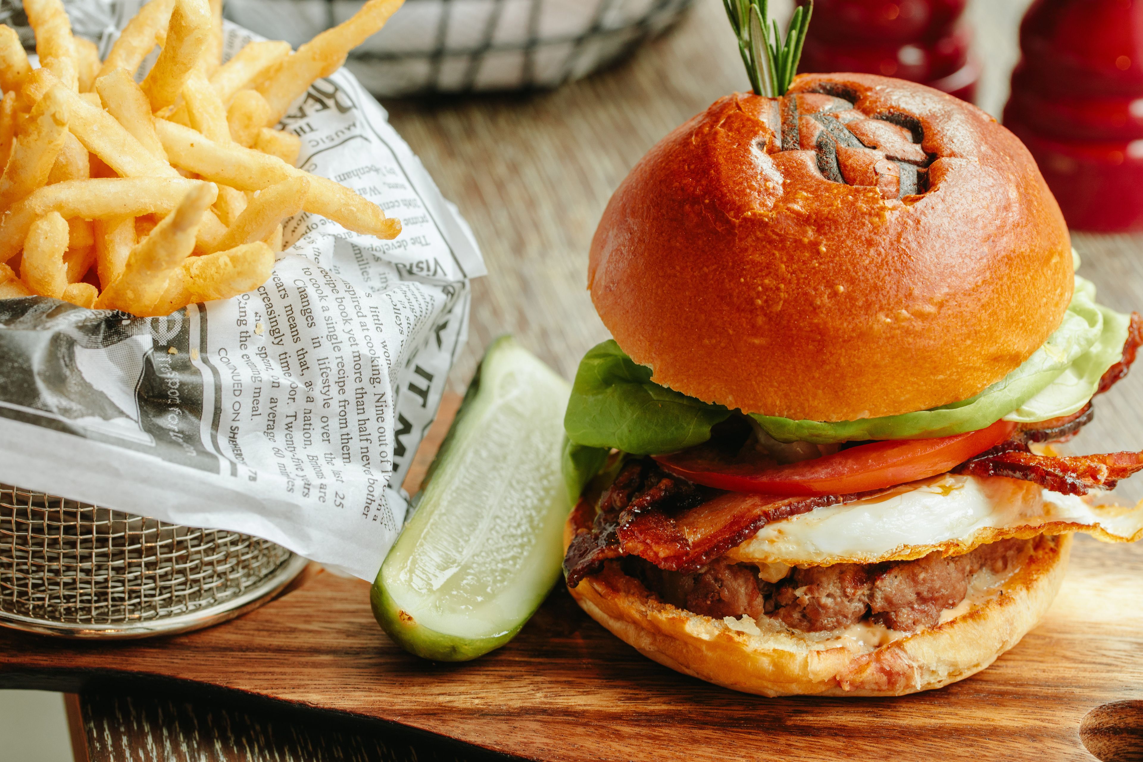 Un hamburger garni de bacon, d’œuf et de laitue, servi dans un pain, accompagné de frites et d’un cornichon.