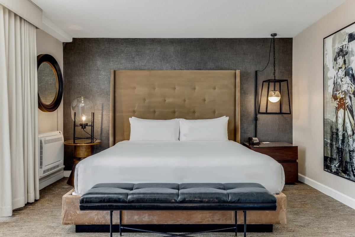 Ein Hotelzimmer mit einem großen Bett, einem Nachttisch mit Lampe, einem Wandbild und einem Fenster mit Vorhängen.