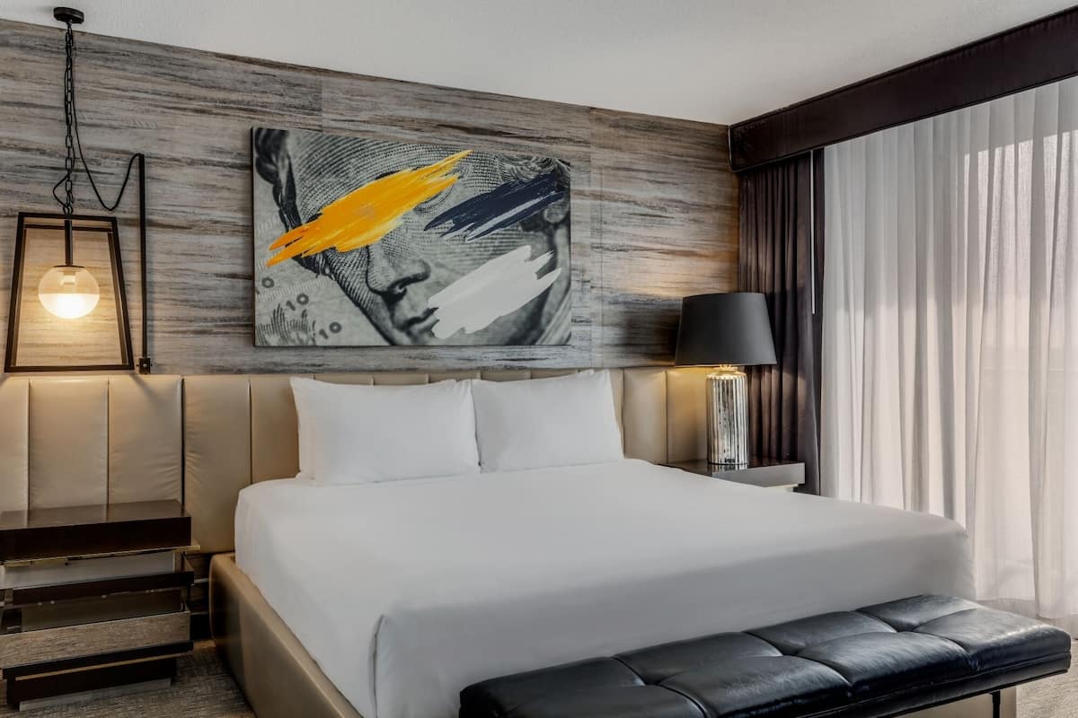 Ein Hotelzimmer mit einem Bett, einer Sitzbank, einem Nachttisch, einer Lampe und einem Wandbild.