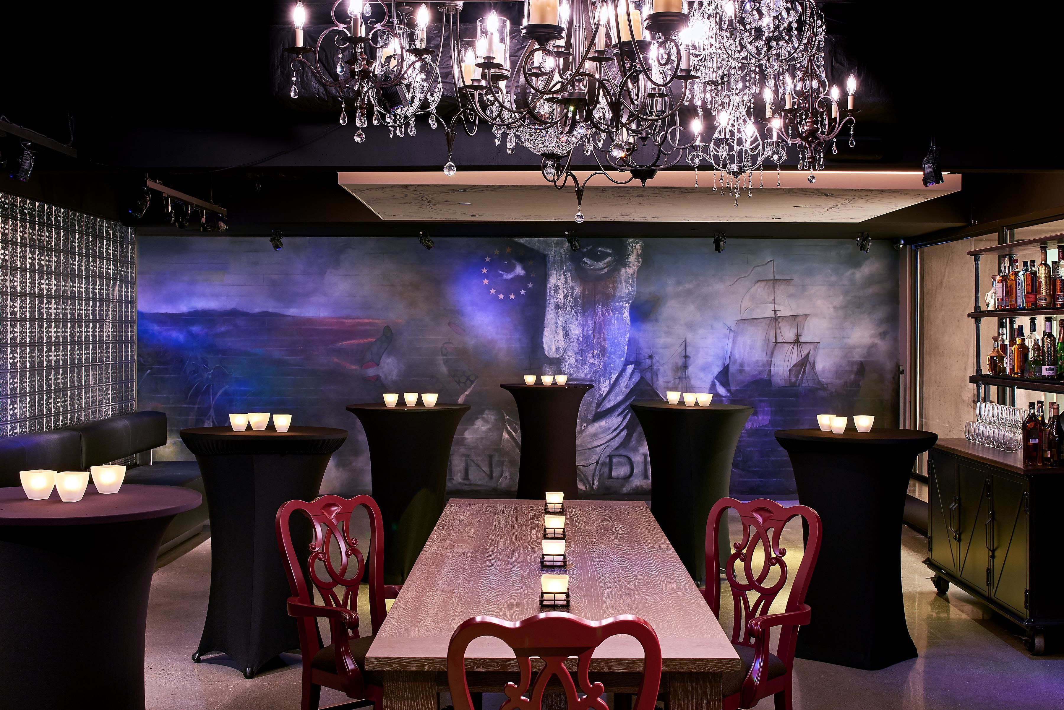 Un área de bar tenuemente iluminada con una mesa larga, sillas rojas y un candelabro.