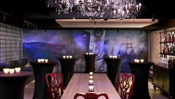Un área de bar tenuemente iluminada con una mesa larga, sillas rojas y un candelabro.