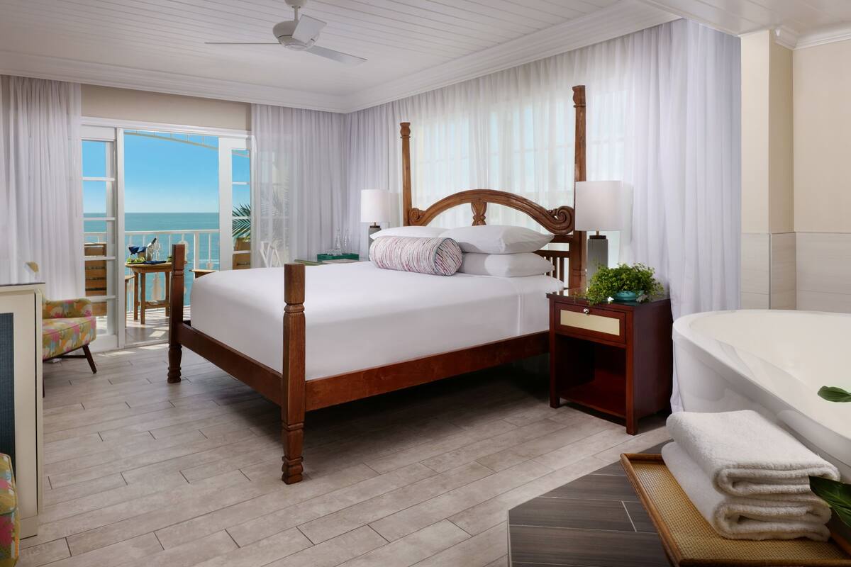 A hotel room with a large bed, a bedside table with a lamp, a bathroom with a bathtub, and a balcony with a view of the ocean.