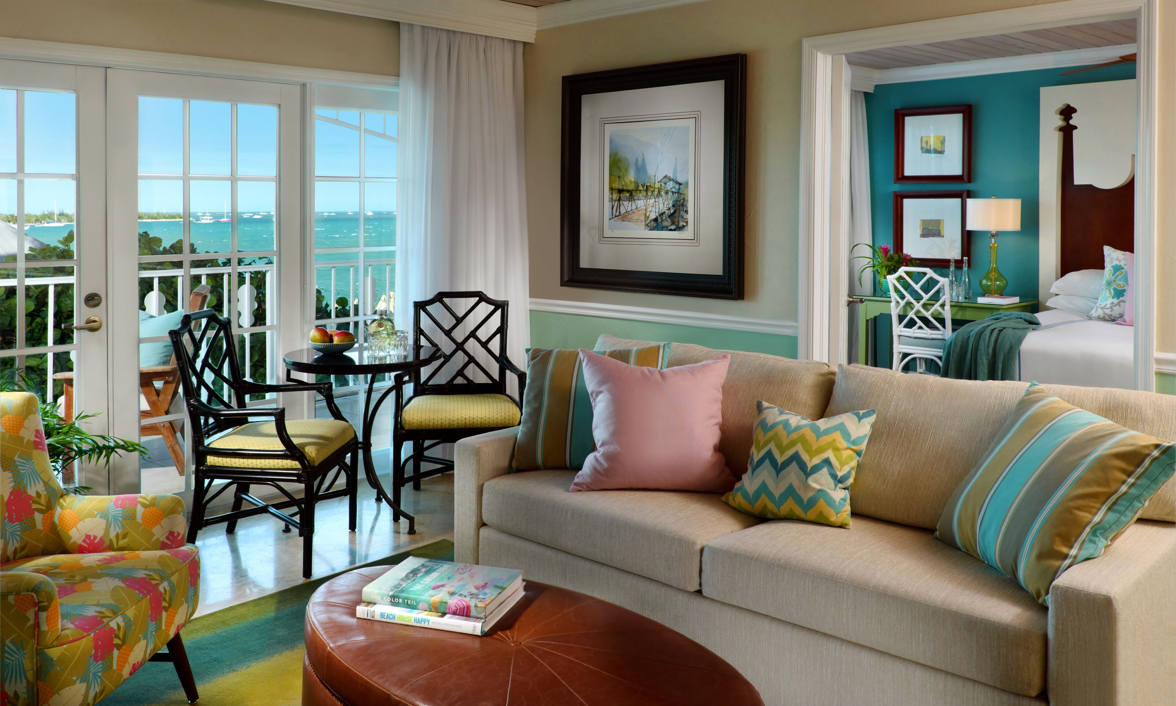 A living room with a beige sofa, a coffee table, and a dining area with chairs and a table. There is a view of the sea through large windows.