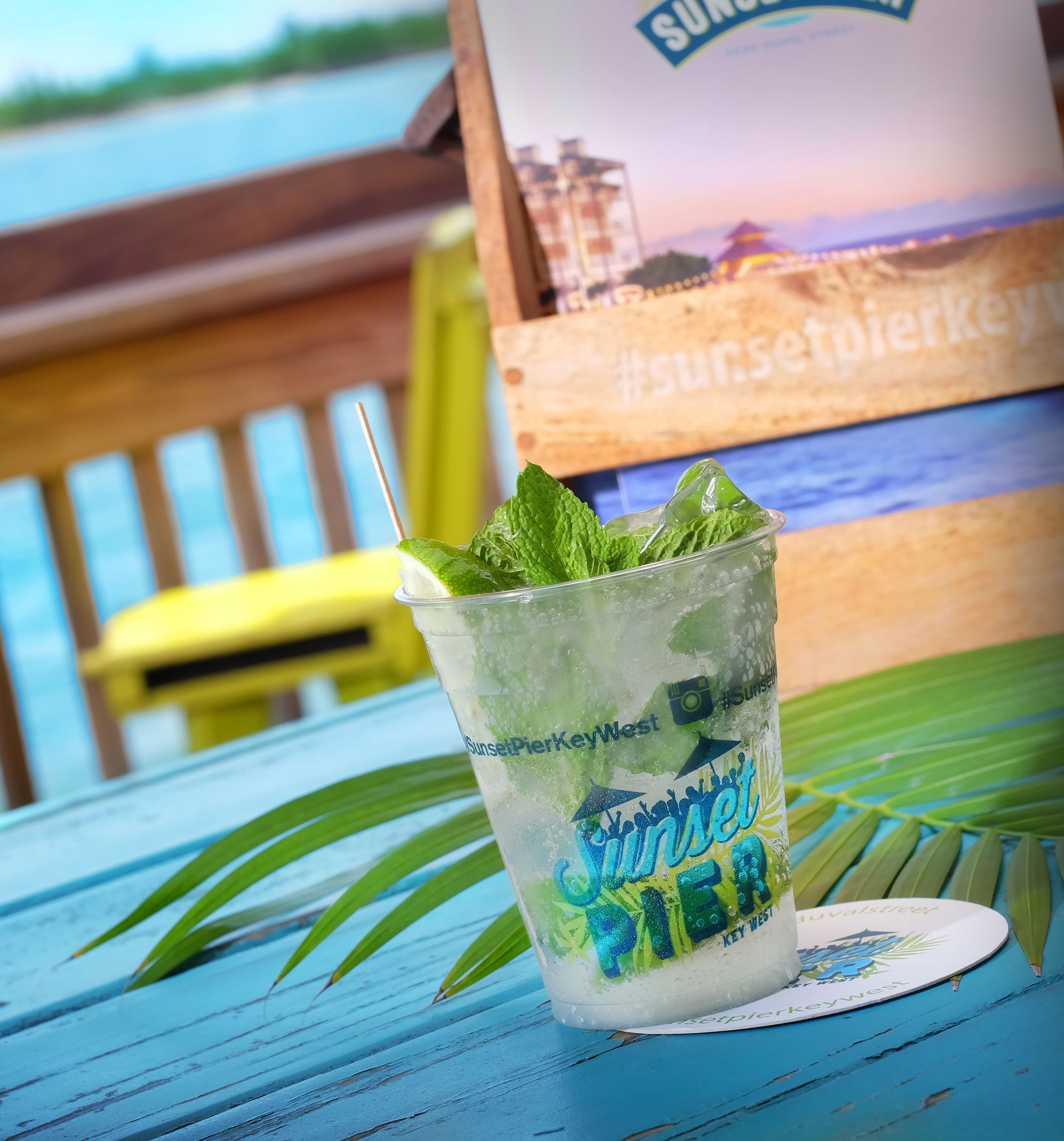 A glass of Sunset Pier Key West cocktail with mint leaves, placed on a blue wooden table with a palm leaf.