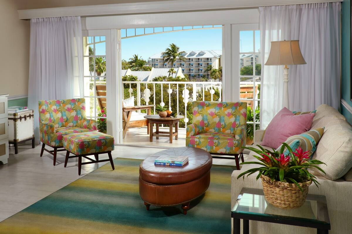 A room with a sofa, two armchairs, a coffee table, a lamp, and a view of a building through large windows.