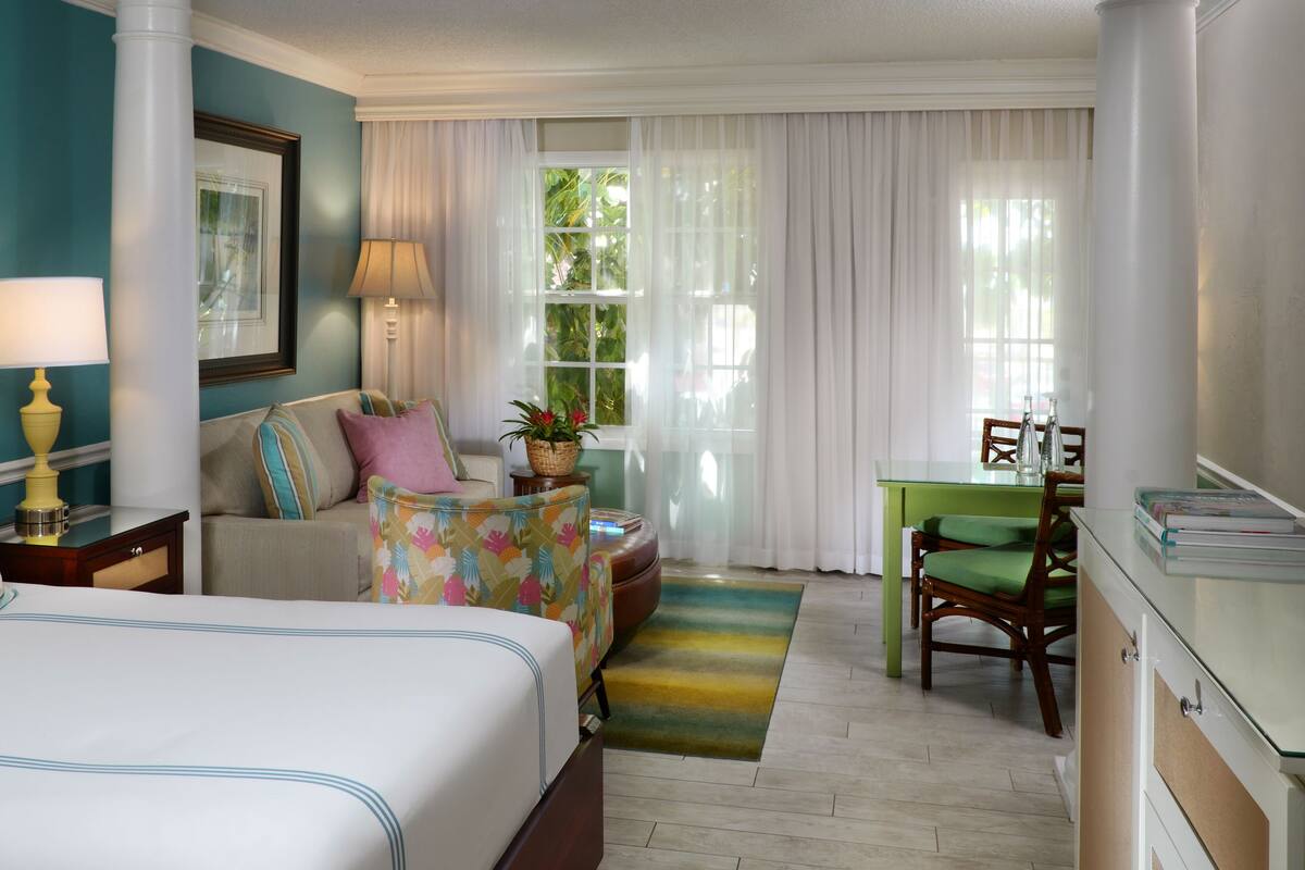 A hotel room with a bed, a sofa, a desk with a chair, a window with curtains, and a plant on the windowsill.