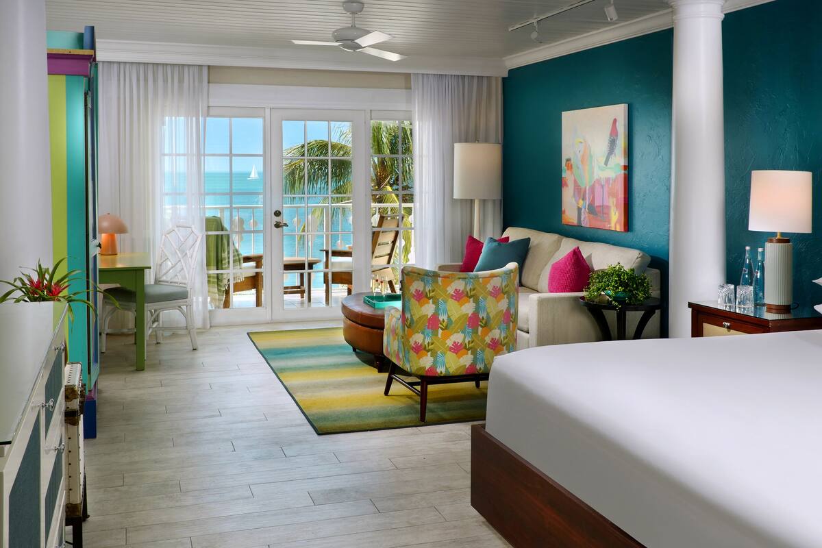 A hotel room with a bed, a sofa, a chair, a table, and a view of the ocean through large windows.