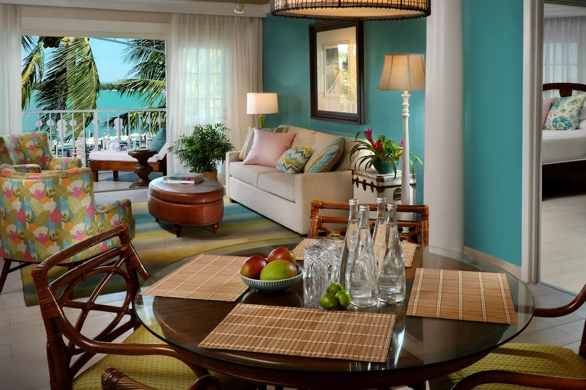 A well-lit living room with a sofa, armchairs, a dining table with placemats, and a view of the outdoors through large windows.