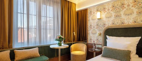A hotel room with a bed, a sofa, a chair, a small table with a vase of flowers, and a wall with floral wallpaper.