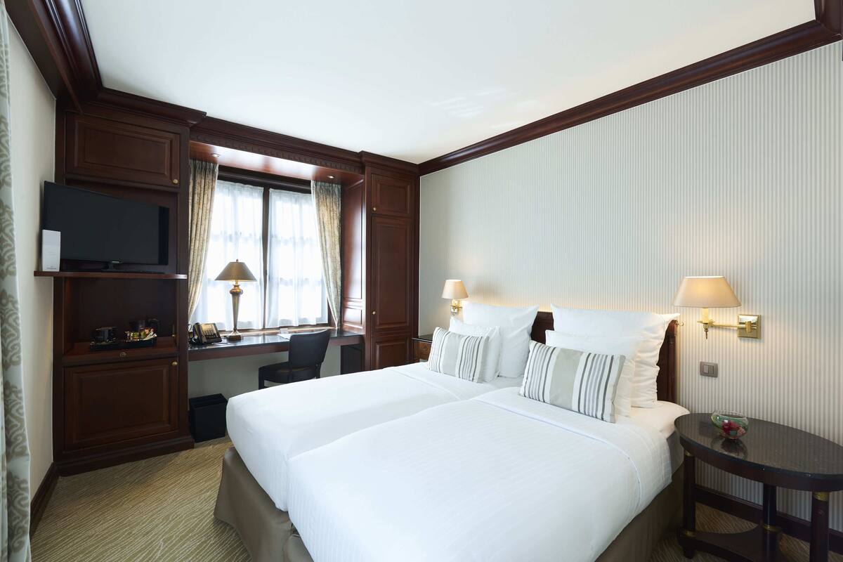 A hotel room with two beds, a desk with a TV, and a window with curtains.