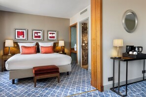Una habitación de hotel con una cama grande, dos mesitas de noche con lámparas, un banco, una pequeña mesa con cafetera y un espejo en la pared.