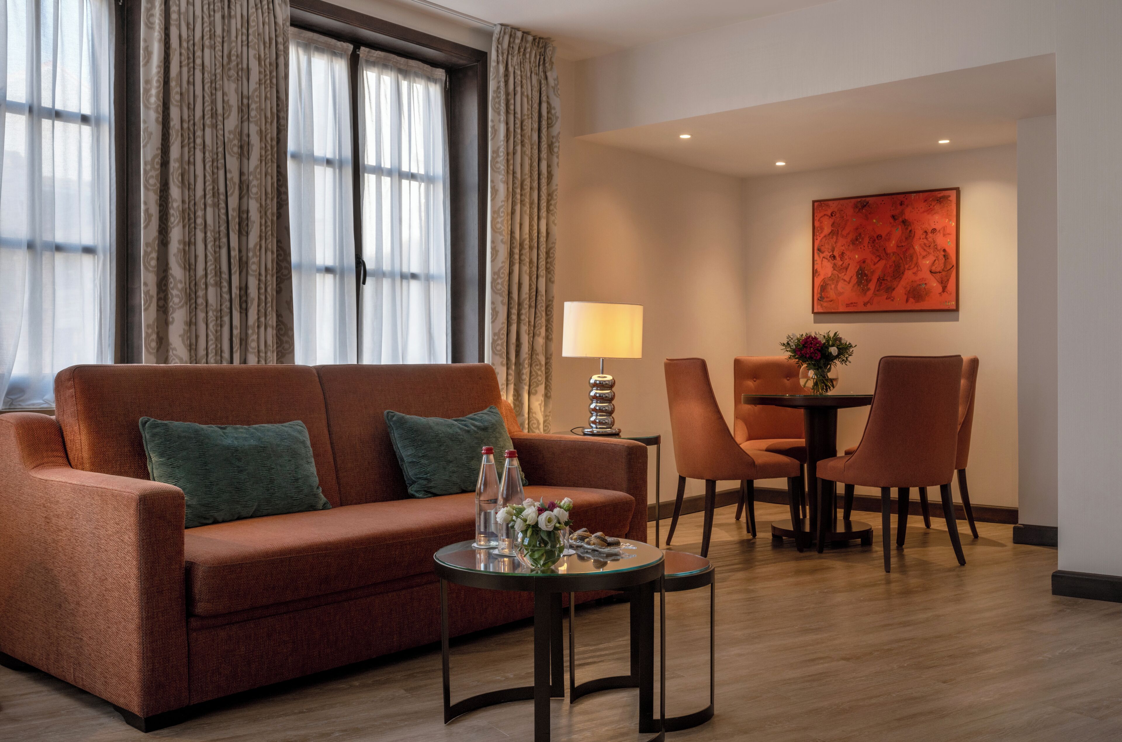 A hotel room with a sofa, a small table with a lamp, a dining table with chairs, and a large window with curtains.