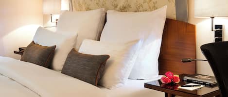 Una cama bien hecha con almohadas blancas y marrones, un cabecero de madera y una mesita de noche con una lámpara, libros y un florero.