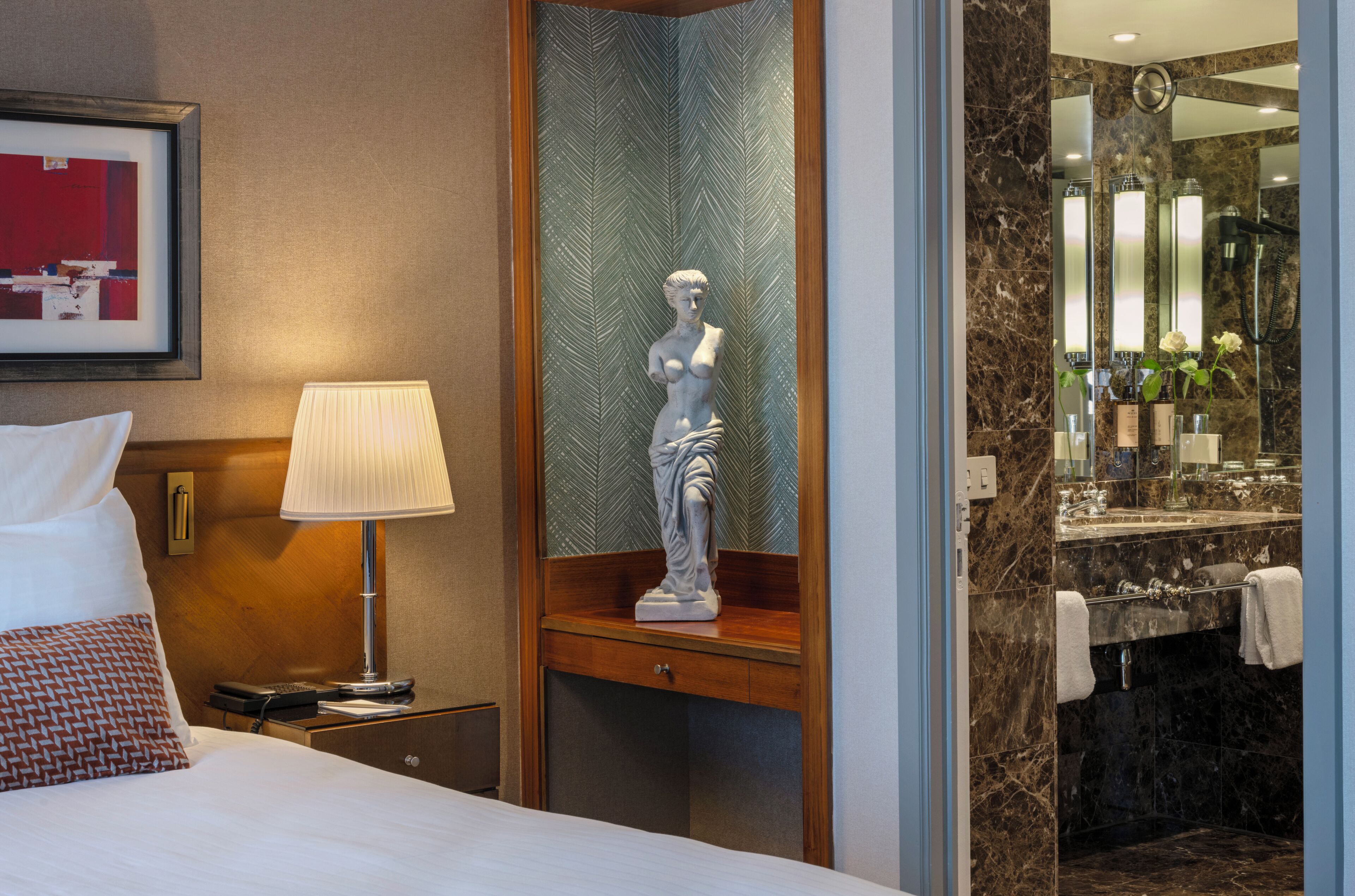 A hotel room with a bed, a nightstand with a lamp, a statue, a bathroom with a mirror, and a shower.
