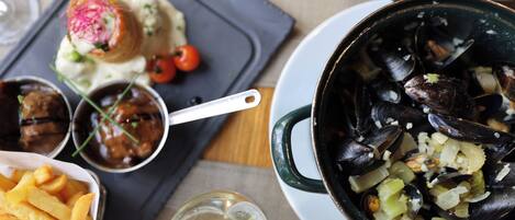 Una mesa de comedor con una variedad de platos, incluyendo mejillones, papas fritas y un guiso de carne, junto con una copa de vino.