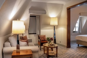 A hotel room with a sofa, armchair, round table, lamps, and a bed.