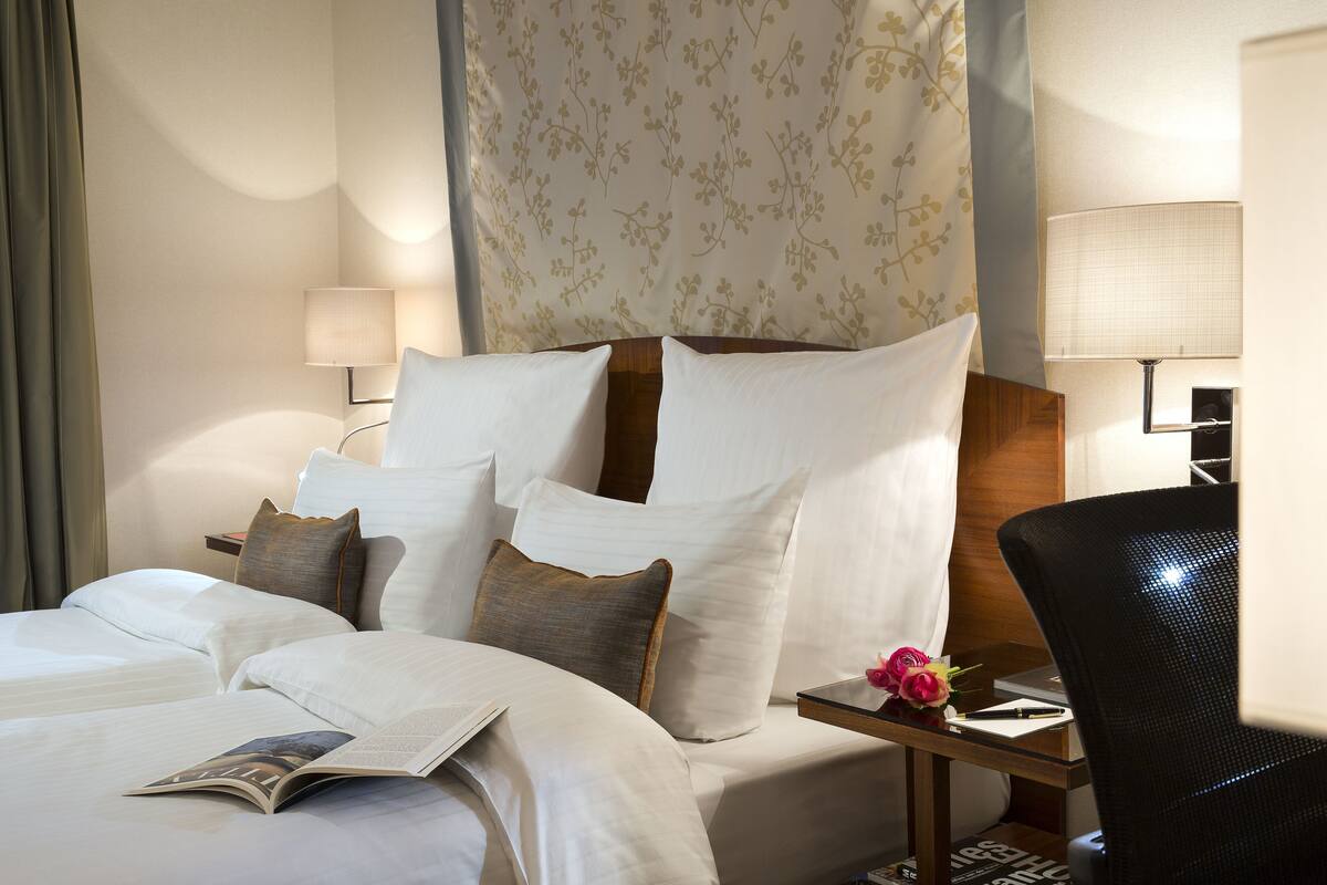 A hotel room with a neatly made bed, a bedside table with a lamp, a chair, and a small table with a flower vase.