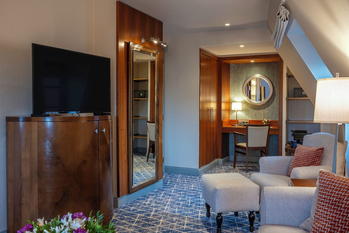 A hotel room with a TV, wooden wardrobe, a dining area with a table and chairs, a mirror, and a lamp.