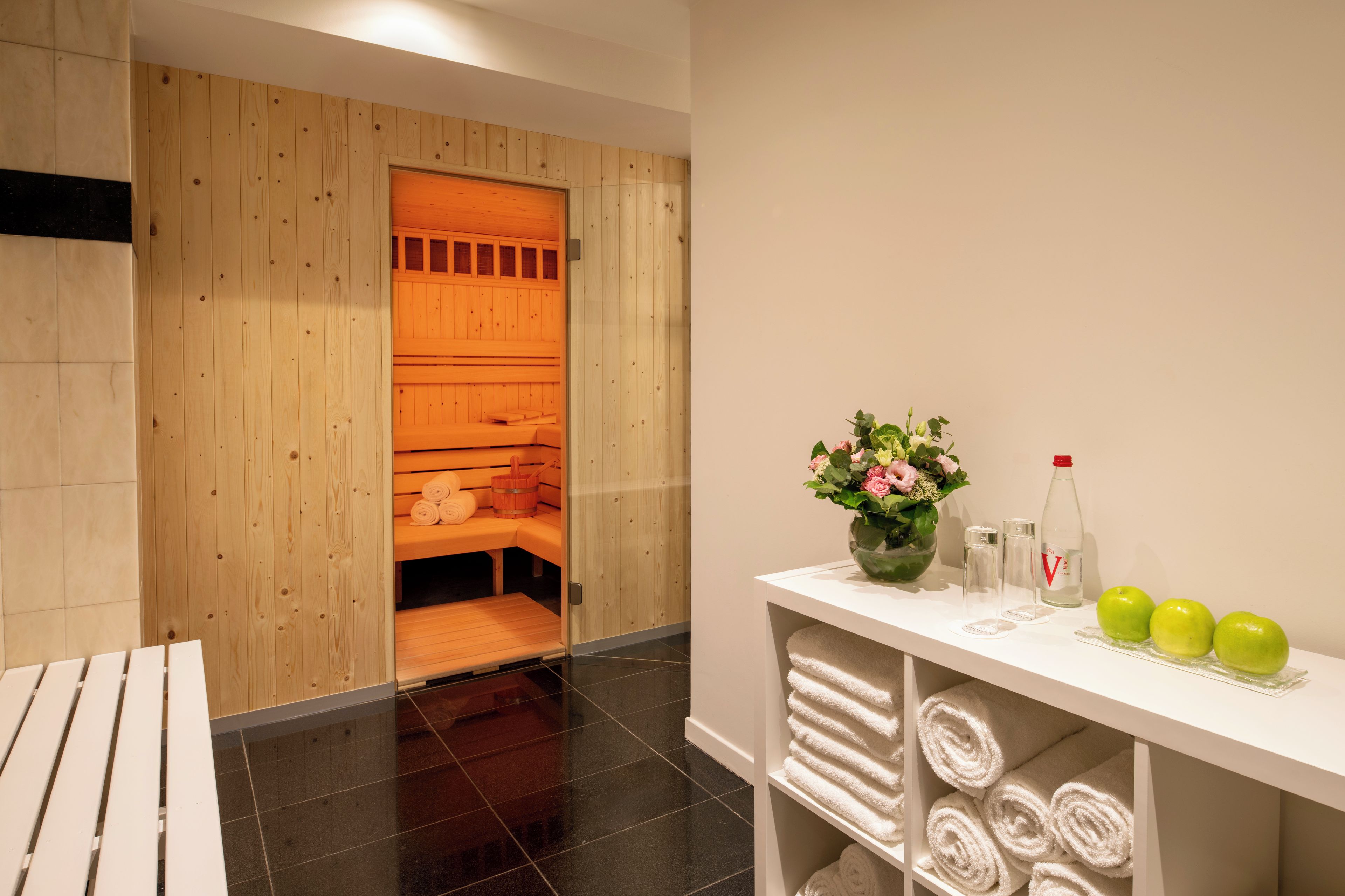 Une salle de sauna avec des bancs en bois et un banc où sont disposées des serviettes. On y trouve une étagère blanche avec des serviettes et un vase rempli de fleurs.