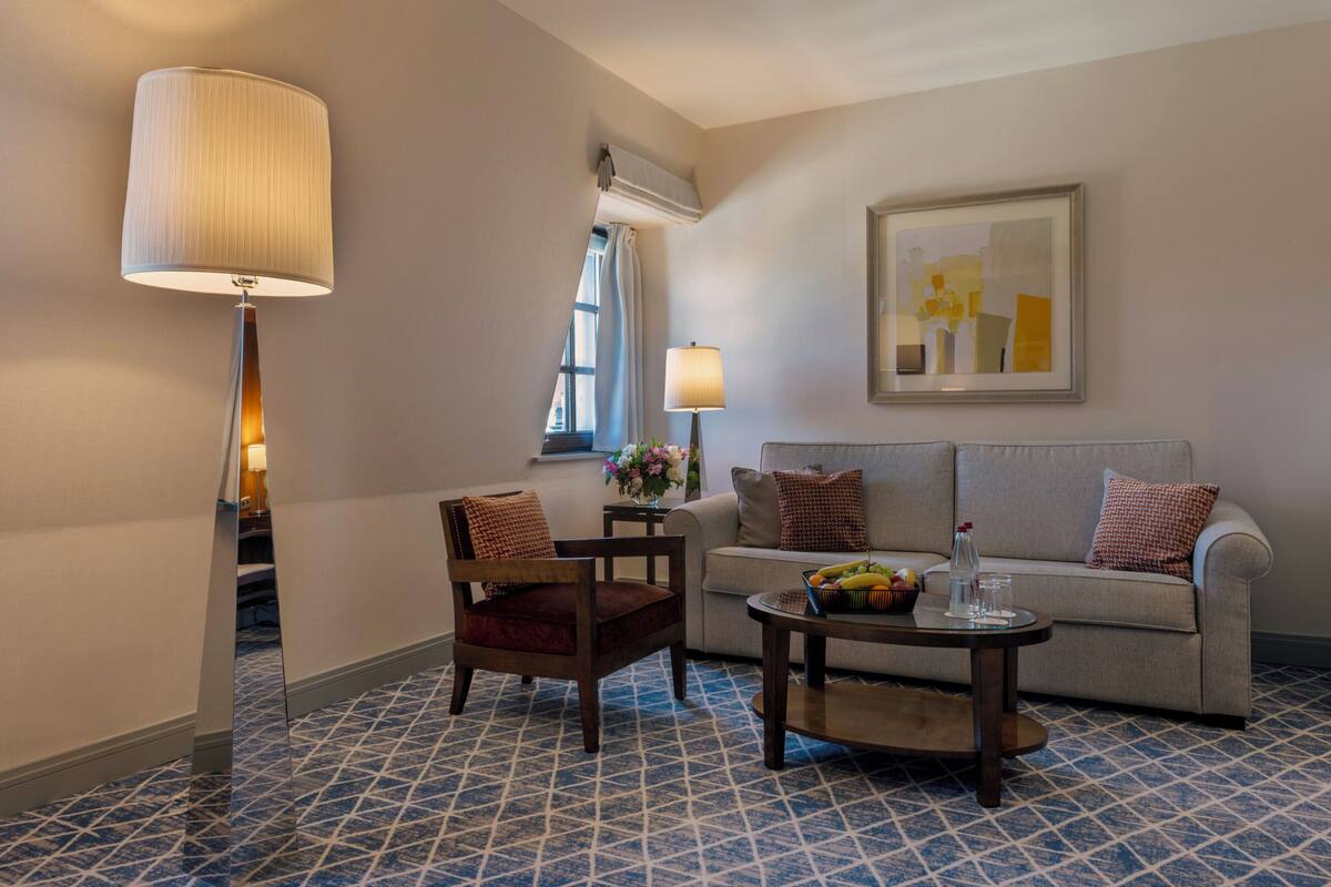 A hotel room with a sofa, armchair, coffee table, lamp, and artwork on the wall.